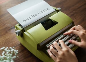 A green typewriter with paper in it that says "marketing." Marketing is very important for video production.